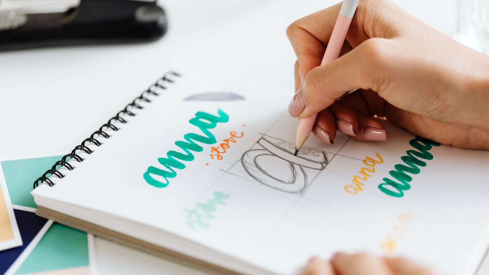 Main qui dessine au crayon les lettres d'un logo dans un carnet, à côté de nuanciers de couleurs